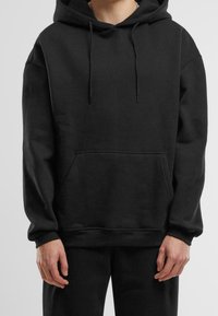 Person trägt einen schlichten schwarzen Kapuzenpullover mit vorderer Kängurutasche und Kordeln und steht vor einem neutralen Hintergrund.