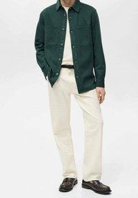 Chemise verte à boutons avec deux poches poitrine, pantalon crème à jambes droites, et chaussures foncées avec détails texturés. Fond uni.