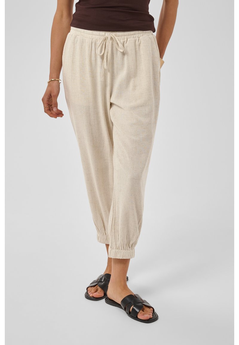 Personne portant un pantalon beige à cordon avec des poignets élastiques et des sandales noires, debout devant un fond clair uni.