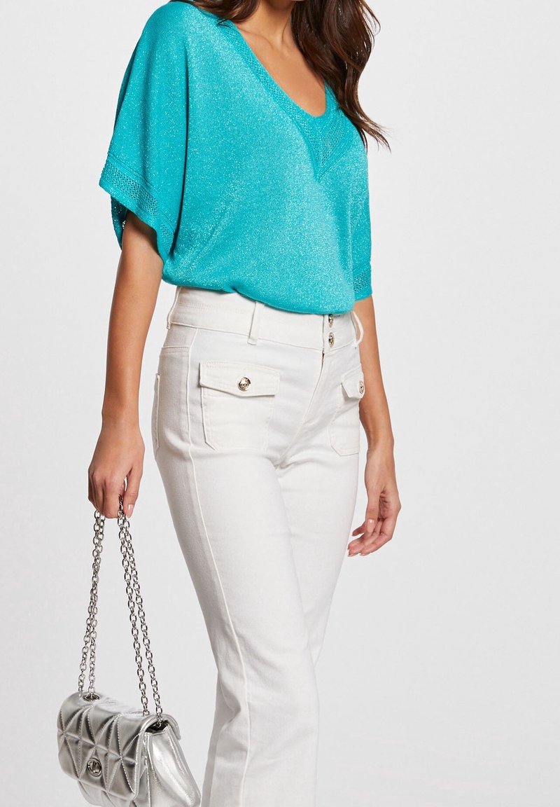 Haut à manches courtes turquoise avec une texture scintillante, associé à un pantalon blanc ajusté avec des poches avant et des boutons dorés. Sac à main argenté.