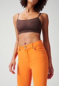 Bruine bralette met spaghettibandjes en logo-band, gecombineerd met strak aansluitende feloranje jeans met knoop en zakdetails.