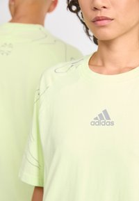 Světle zelené bavlněné tričko s šedým logem Adidas a abstraktními liniovými vzory. Krátké rukávy a kulatý výstřih.