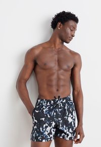 Schwarze Shorts mit einem abstrakten blauen und weißen Muster, ausgestattet mit einem elastischen Bund und einer Kordel. Das Modell ist oberkörperfrei.