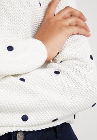 Hand auf dem gegenüberliegenden Arm ruhend, trägt einen weißen Strickpullover mit schwarzen Punkten und eine dunkelblaue Jeans.