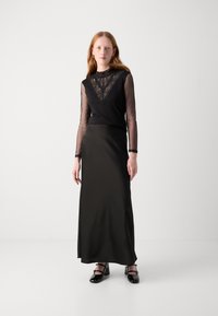 Schwarzes langärmeliges Mesh-Oberteil mit Spitzenverzierungen, kombiniert mit einem schwarzen satin Maxirock. Die Schuhe sind mit geschlossenen schwarzen Absätzen mit Riemen ausgestattet.