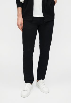 Mand iført sorte slim-fit bukser, hvide sneakers og en delvist lynlåst sort jakke over en hvid skjorte, stående mod en ensfarvet baggrund.