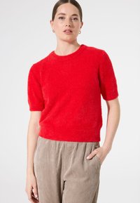Roter kurzärmeliger Pullover aus flauschigem Material mit rundem Halsausschnitt, kombiniert mit hoch taillierten Cordhosen in einem neutralen Farbton.