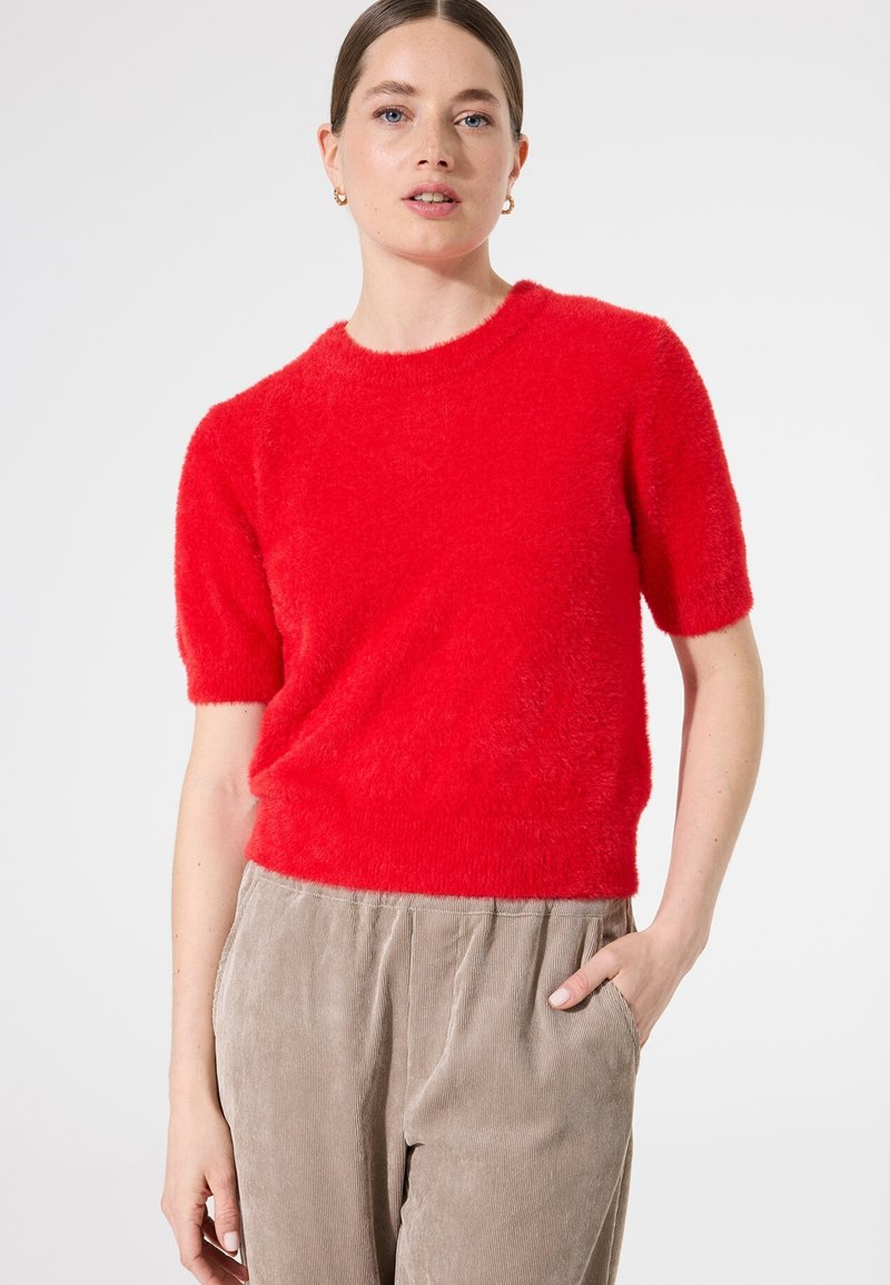 Roter kurzärmeliger Pullover aus flauschigem Material mit rundem Halsausschnitt, kombiniert mit hoch taillierten Cordhosen in einem neutralen Farbton.