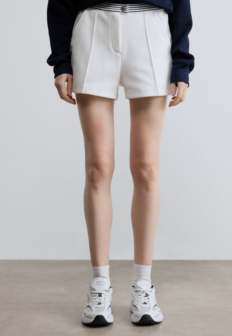 Short blanc plissé avec une ceinture rayée, fabriqué en matériau doux, assorti à des chaussures de sport blanches et des chaussettes jusqu'à la cheville.
