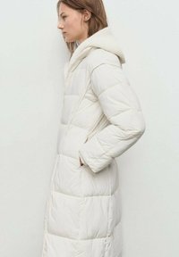 Joven mujer vestida con un abrigo de invierno largo, blanco, acolchado y con capucha, con las manos en los bolsillos, de perfil a la izquierda contra un fondo claro y simple.