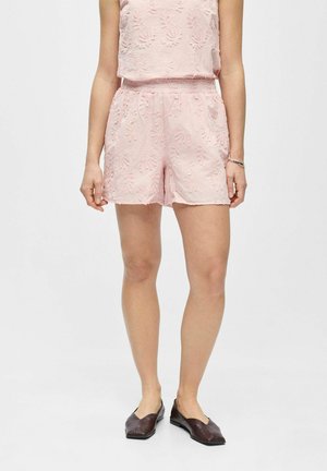 Žena nosi svetloružové kvetované textúrové šortky a zodpovedajúci bezrukávový top, spojené s tmavohnedými plochými topánkami, stojaca na bielom pozadí.