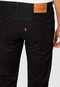 Jeans de mezclilla negra con una superficie texturizada, que cuentan con bolsillos traseros, un parche de cuero marrón y una etiqueta roja en la parte trasera.