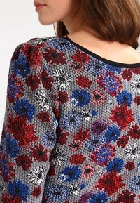 Blouse met bloemenpatroon, met een getextureerde grijze achtergrond en rode, blauwe en witte bloemen. Ronde halslijn met zwarte bies.