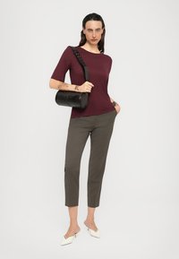 Woman wearing a burgundy top, gray cropped pants, white slingback heels, and carrying a black cylindrical shoulder bag, standing against a plain background.