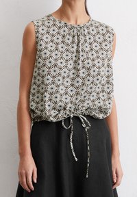 Beige-mustavalkoinen, kukkakuvioinen hihaton paita, jossa on poimutettu pääntie ja säädettävä vyötärönauha, yhdistetty mustaan hameeseen.