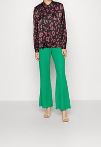 Femme portant une blouse noire à manches longues avec un imprimé léopard rouge et un pantalon large texturé vert vif avec des talons ouverts beiges.