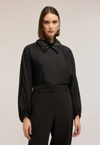 Blusa negra con diseño de manga abullonada, con un gran cuello adornado con tachuelas blancas. Combinada con pantalones negros de talle alto.