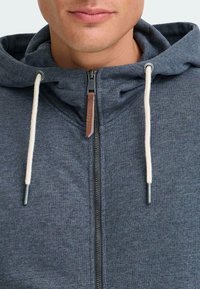 Šedá mikina na zips s predným zipsom, béžovými šnúrkami a jemnou textúrou. Vyznačuje sa obrysovým švom pozdĺž goliera a kapucne.