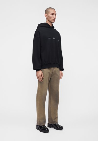 Homme portant un sweat à capuche noir avec des lettres, un pantalon marron et des chaussures à plateforme noires, debout devant un fond gris clair uni.
