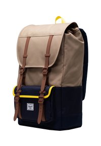 Kanvasreppu ruskeassa ja tummansinisessä värissä, jossa on ruskeat nahkaremmit, keltainen reunus ja läppäsulku. Suunniteltu etutaskulla säilytystä varten.