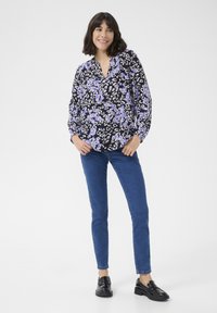 Blouse noire à imprime léopard violet et blanc, décolleté en V, manches longues ; associée à un jean bleu ajusté et des mocassins noirs.