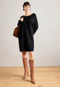 Μαύρο oversized πλεκτό φόρεμα με φαρδύ ντεκολτέ, σε συνδυασμό με καφέ ψηλοτάκουνα μποτάκια μέχρι το γόνατο. Το μοντέλο κρατά μια καφέ σουέτ τσάντα ώμου.
