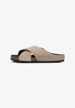 Suede slide sandal in light beige featuring an asymmetrical cross-strap design, cork footbed, and textured black rubber outsole.