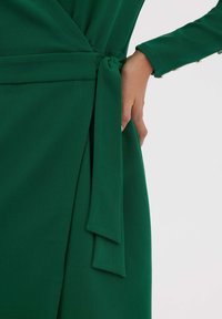 Robe verte enroulée avec une taille attachée, manches longues et texture de tissu lisse, présentant un éclat subtil et des lignes épurées.