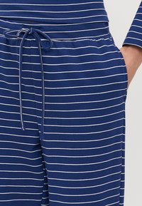 Modré pruhované pyžamo s bielymi horizontálnymi pruhmi, obsahujúce elastický pás a šnúrku na zaviazanie, mäkký materiál a bočné vrecká.