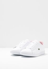 Sneakers bianchi Lacoste con imbottitura rosa, caratterizzati da un logo coccodrillo verde, esposti su uno sfondo bianco semplice.