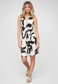 Mouwloze jurk in crème met zwarte bladerenpatronen, voorzien van een geknoopte taille en een relaxte pasvorm. Gemaakt met lichtbruine sandalen.