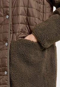 Gequiltte bruine buitenlaag met een zachte, gestructureerde fleece zak. Bevat drukknoopjes en een aardse kleurenschema.