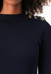 Top in maglia a coste di colore navy con collo alto e bottoni decorativi dorati sulla spalla. Tessuto testurizzato con un design aderente.
