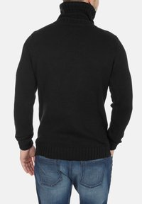 Suéter de punto negro con un cuello alto acanalado y borde acanalado, que presenta un patrón de tejido texturizado. Combinado con pantalones vaqueros de mezclilla azul.