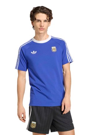 Jeune homme portant un maillot de football bleu Adidas Argentina et un short noir avec des logos, debout devant un fond blanc.