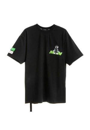 Joven mujer de pie, con una camiseta negra de gran tamaño con gráfico y pantalones negros a juego con detalles verdes, zapatos negros texturizados.