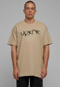 Beige oversized cotton t-shirt with short sleeves and a round neckline; features a black graphic text design across the front.