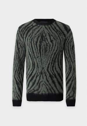 Homme debout portant un pull à motifs verts et noirs, un pantalon noir à jambes larges et des chaussures en cuir noires sur un fond gris clair.