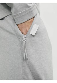Sudadera con cremallera de color gris, fabricada con una suave mezcla de algodón, con costuras visibles y acentos en gris en los cordones con puntas de plástico. Detalle del bolsillo lateral visible.