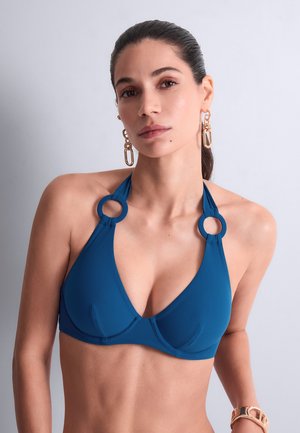 Femme aux longs cheveux foncés portant un haut de bikini bleu dos nu, des boucles d’oreilles en chaîne dorée et un bracelet en or sur un fond clair uni.