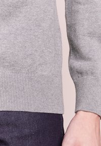 Nahaufnahme einer Person, die einen hellgrauen gestrickten Pullover und eine dunkelblaue Jeans trägt, wobei der Ärmel und der Taillenbereich sichtbar sind.