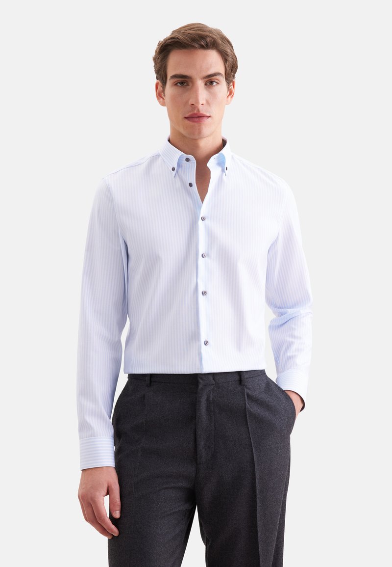 Jeune homme portant une chemise à rayures bleu clair boutonnée et un pantalon gris foncé, debout avec une main dans la poche sur fond blanc.