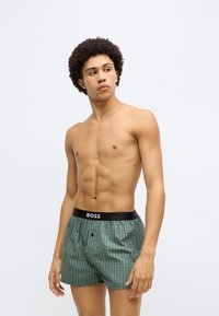 Zöld kockás boxeralsó fekete derékpánttal, amelyen "BOSS" logó található, puha anyagból készült. A dizájn gombos és kényelmes szabású.