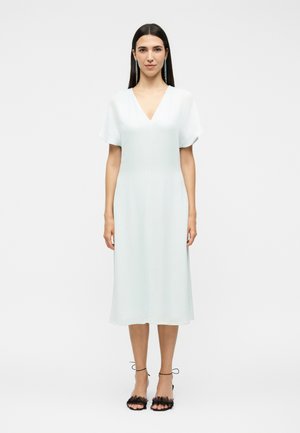 Γυναίκα που στέκεται με λευκό midi φόρεμα με κοντά μανίκια και V-λαιμόκοψη, φορώντας μαύρα ψηλοτάκουνα πέδιλα με λουράκια και μακριά σκουλαρίκια, σε απλό φόντο.