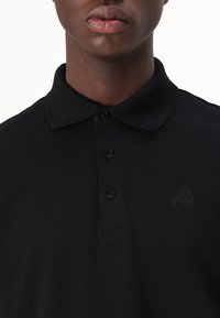 Black polo shirt in cotton material with a classic collar, three buttons, and a subtle embroidered logo on the chest. Smooth texture.