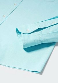 Hellblaues Hemd mit Knopfleiste und gefaltetem Ärmelabschluss, ausgestattet mit weißen Knöpfen und einer glatten, weichen Textur. Keine Muster oder zusätzlichen Verzierungen.