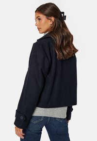 Námornicky modrá zkrácená bunda z vlny se sklopeným límcem a knoflíkovými manžetami, nošená přes světle šedý žebrovaný svetr, v kombinaci s tmavými džíny.