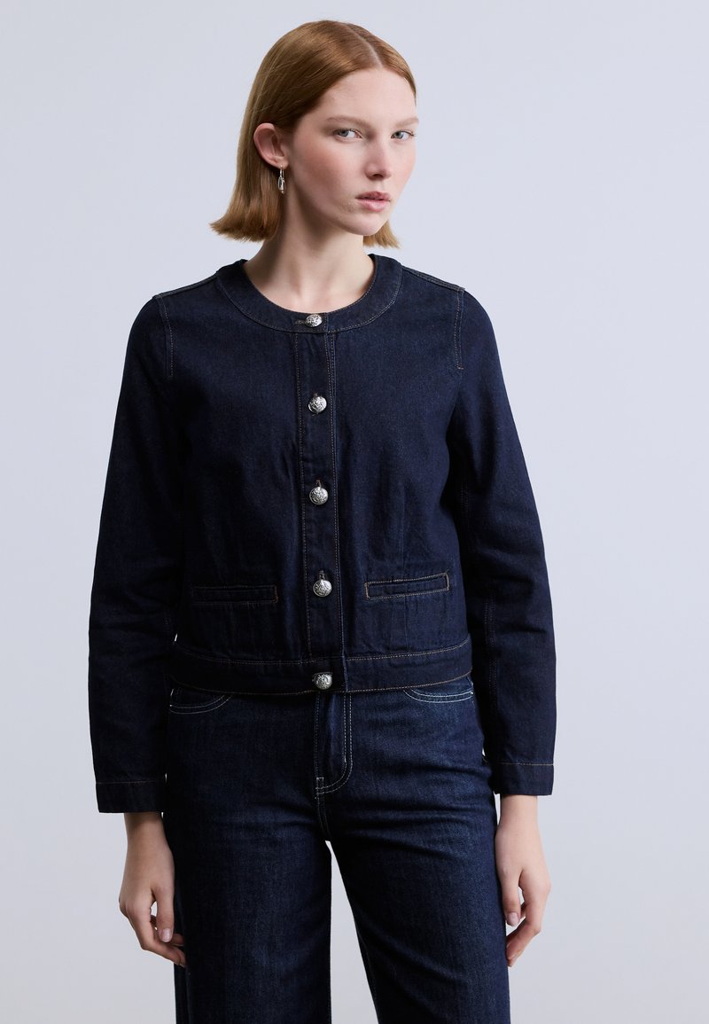 Σκούρο τζιν μπουφάν με στρογγυλή λαιμόκοψη, ασημί κουμπιά, δύο μπροστινές τσέπες και ελάχιστες ραφές. Συνδυασμένο με τζιν παντελόνι στο ίδιο χρώμα.