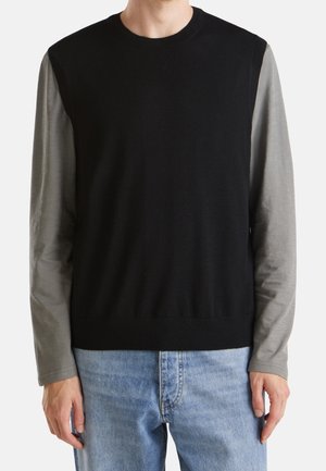 Homme portant un pull sans manches noir par-dessus une chemise grise à manches longues et un jean bleu clair, visible du cou jusqu'aux hanches.