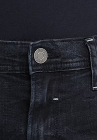 Sorte denimjeans med en metallknapp med "BLEND" gravering, hvite sømmer og en glatt tekstur. Nærbilde av midjeområdet.